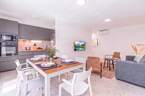Une cuisine moderne dotée d’appareils électroménagers intégrés, un coin repas avec une table en bois et des chaises blanches, et un salon avec un canapé et un tableau au mur.
