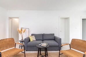 A modern living room with a grey sofa, two wicker chairs, a small round table with books, and a vase with dried plants.