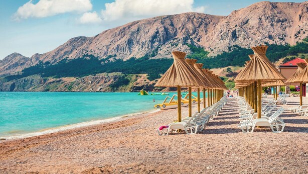 A beach with thatched umbrellas and sun loungers, a rocky mountain in the background, and a clear blue sea. - Pool Apartment Sapore Di Mare 1, Baška, Croatia (Baška)