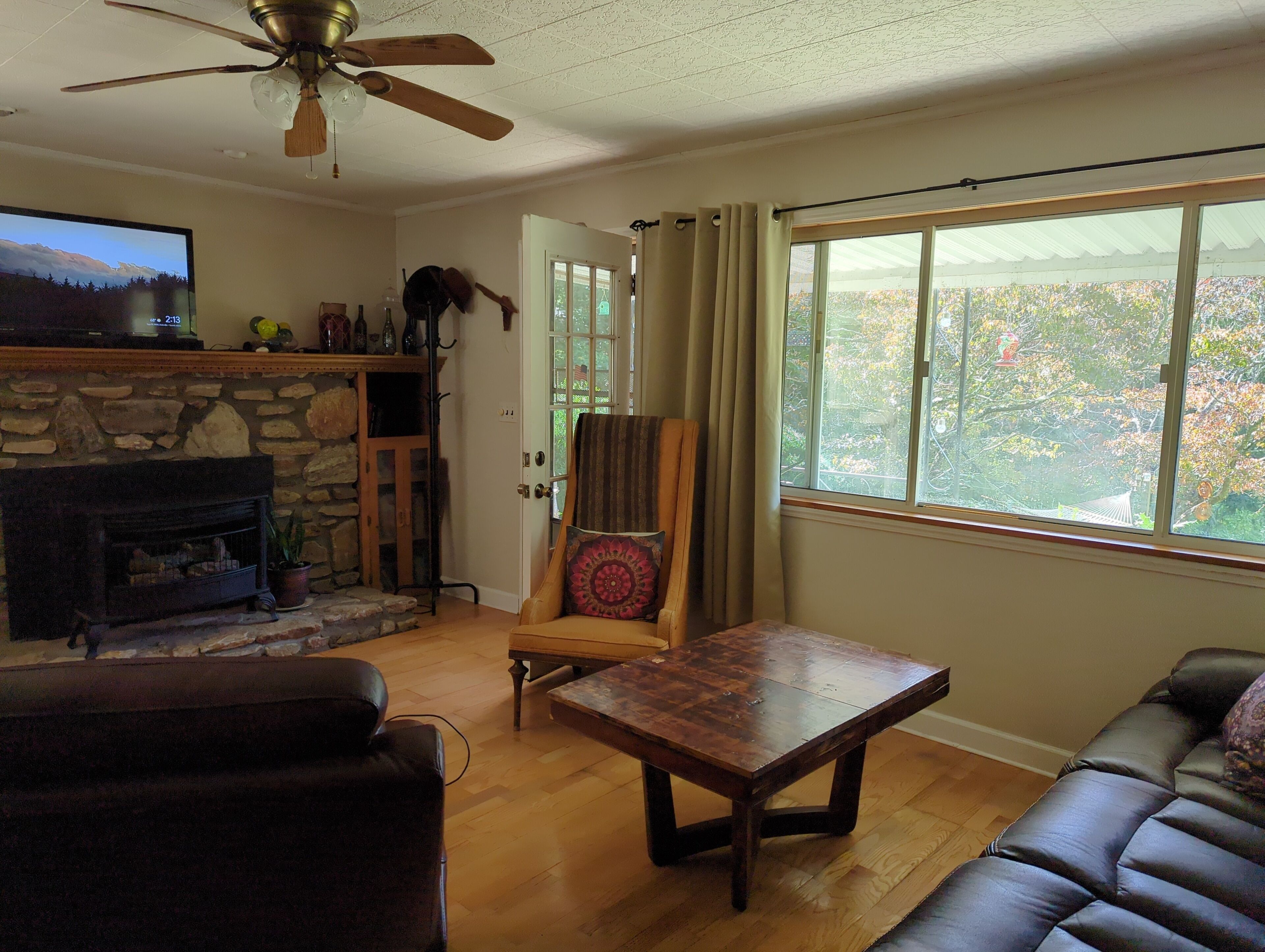 A living room with a stone fireplace, a television, a ceiling fan, a large window with a view, and a wooden coffee table.