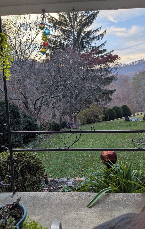 Vista desde un balcón con barandilla, un jardín con diversas plantas, un árbol y una edificación con colinas a lo lejos.