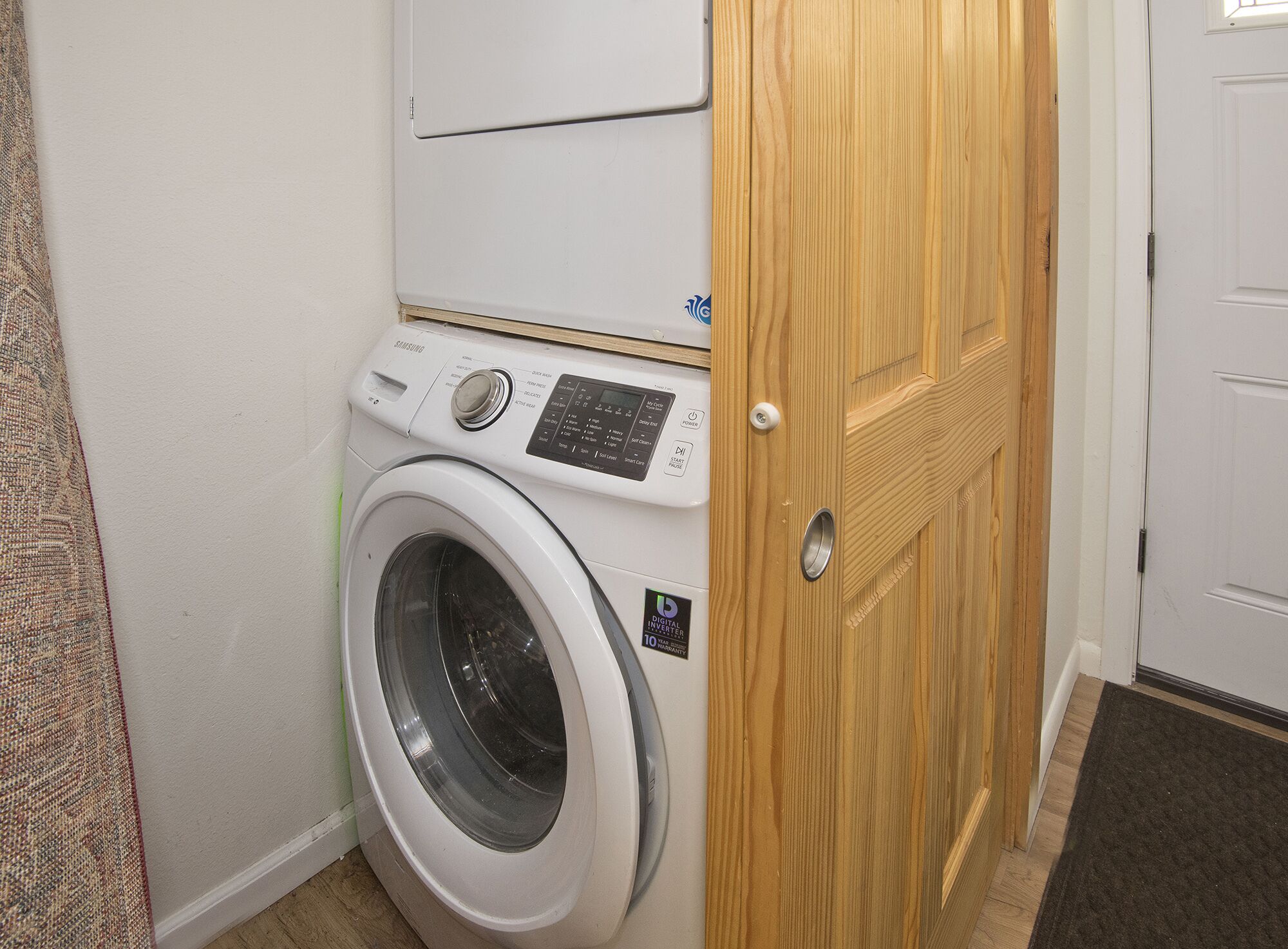 Eine weiße Waschmaschine mit Bedienfeld und Digital-Inverter-Aufkleber, neben einer Holztür in einem Raum mit Teppichboden.