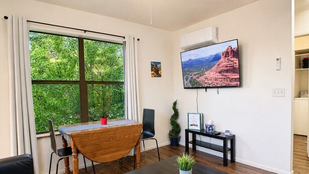 A modern living room with a dining table, a TV mounted on the wall, and a large window with a view of greenery. - Grand Canyon Basecamp with EV (Williams)
