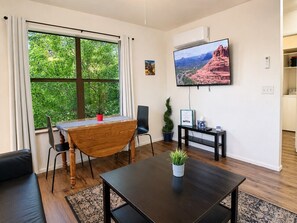 Uma sala de estar moderna com uma mesa de jantar, uma TV fixada na parede e uma grande janela com vista para o verde.