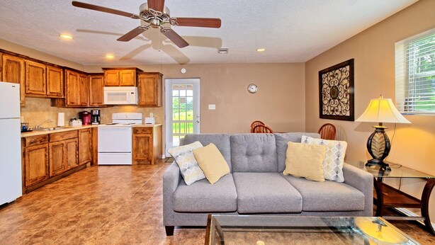 A compact living space with a kitchenette, a grey sofa, a glass coffee table, and a ceiling fan.