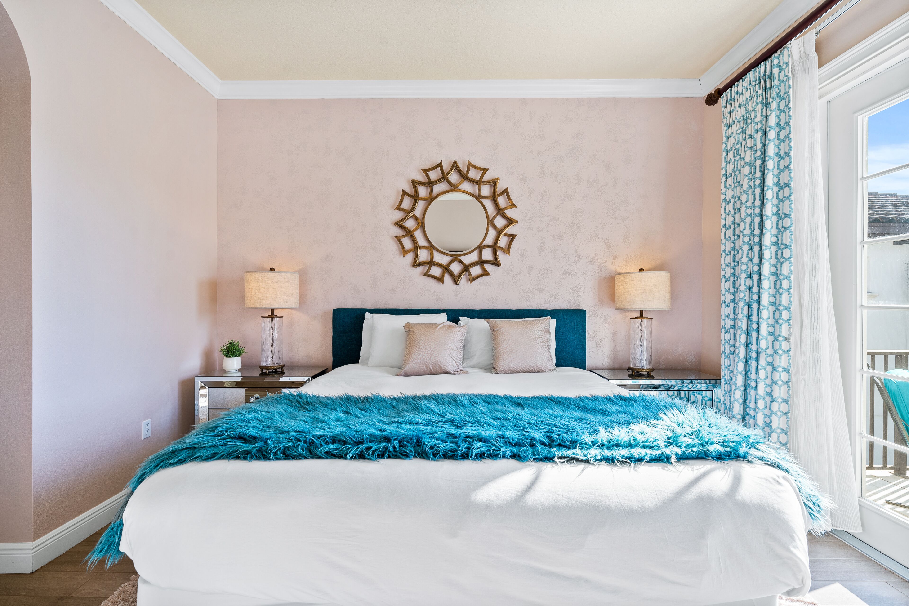 A bedroom with a large bed, two bedside tables with lamps, a decorative mirror on the wall, and a view of the outdoors through a window with curtains.