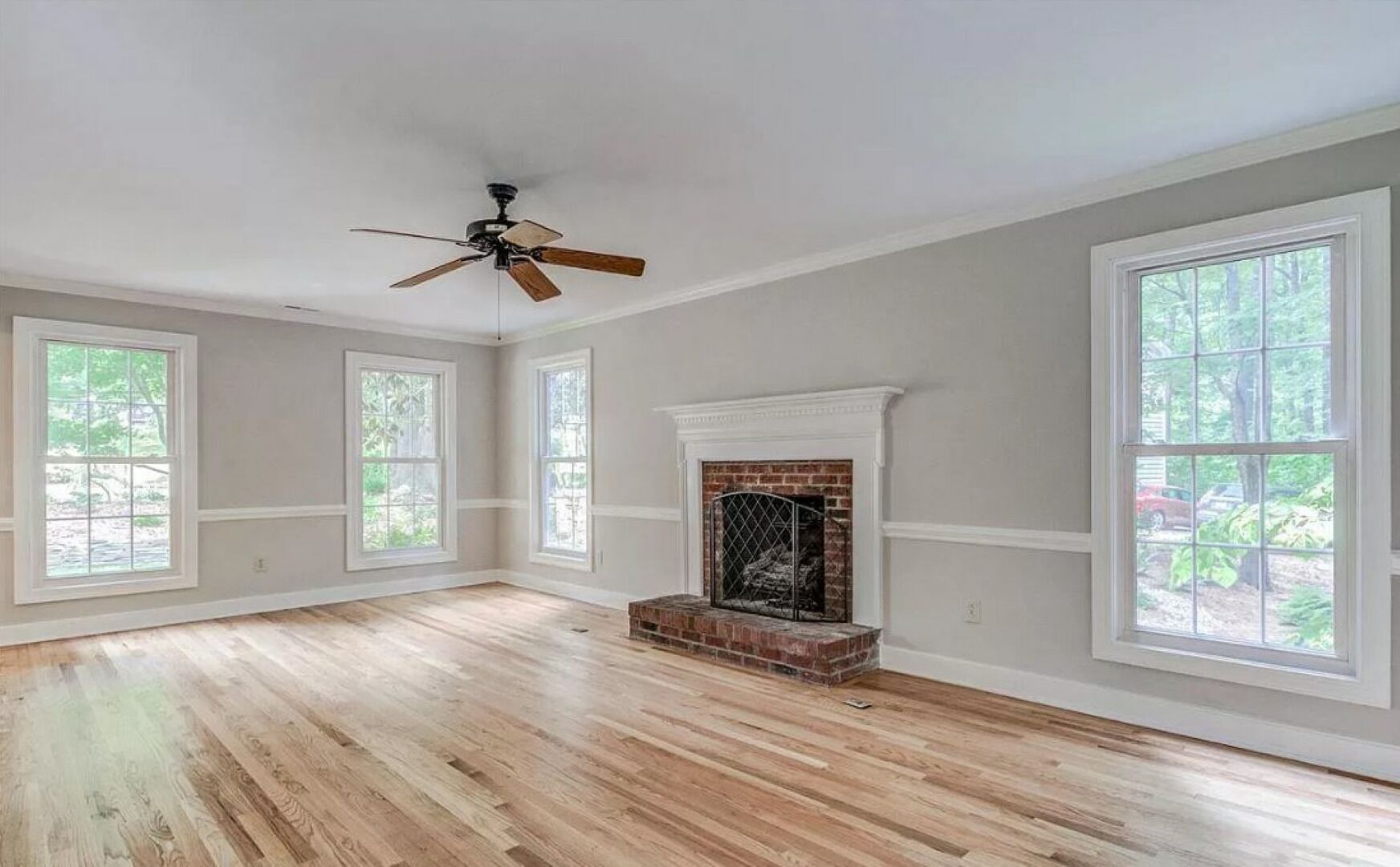 A spacious room with hardwood flooring, large windows, a fireplace, and a ceiling fan.