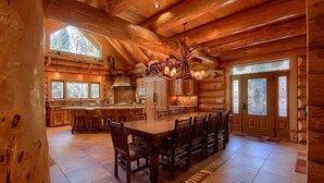A log cabin interior with a large dining table, wooden chairs, a chandelier, and a kitchen area.
