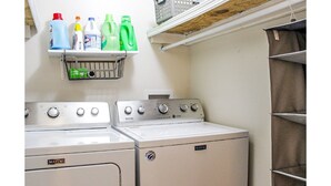 Un área de lavandería con dos lavadoras Maytag, una estantería con botellas de detergente y un tendedero metálico.