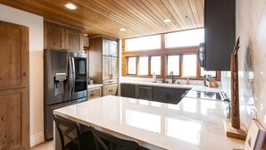 A modern kitchen with wooden cabinets, a stainless steel refrigerator, a large island with bar stools, and a window providing natural light. - Stunning Eastern Sierra Retreat: 3 Bed+Loft/3 Baths, Spacious, Pet-Friendly (Mammoth Lakes)