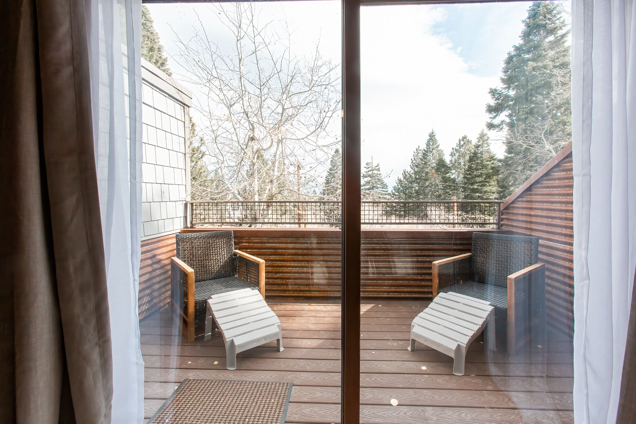 A wooden deck with two wicker chairs and a bench, overlooking a fenced area with trees.