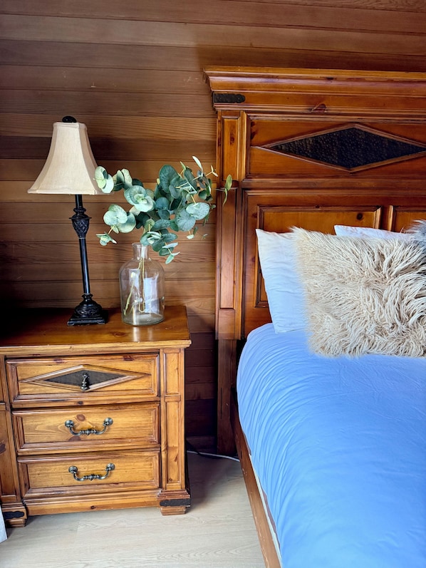 A wooden nightstand with a lamp, a vase with green leaves, and a bed with a blue bedspread and a fluffy pillow. - Stunning Eastern Sierra Retreat: 3 Bed+Loft/3 Baths, Spacious, Pet-Friendly (Mammoth Lakes)
