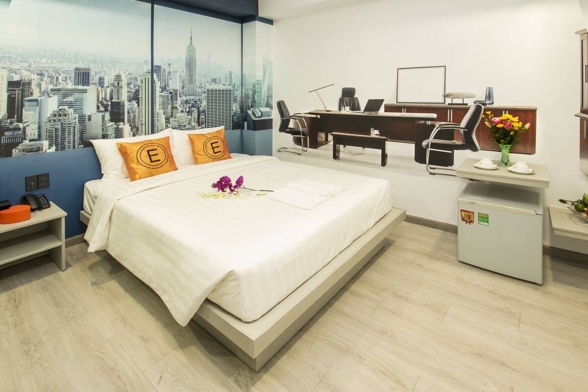 A hotel room with a bed, a desk, a chair, a small refrigerator, and a cityscape view.