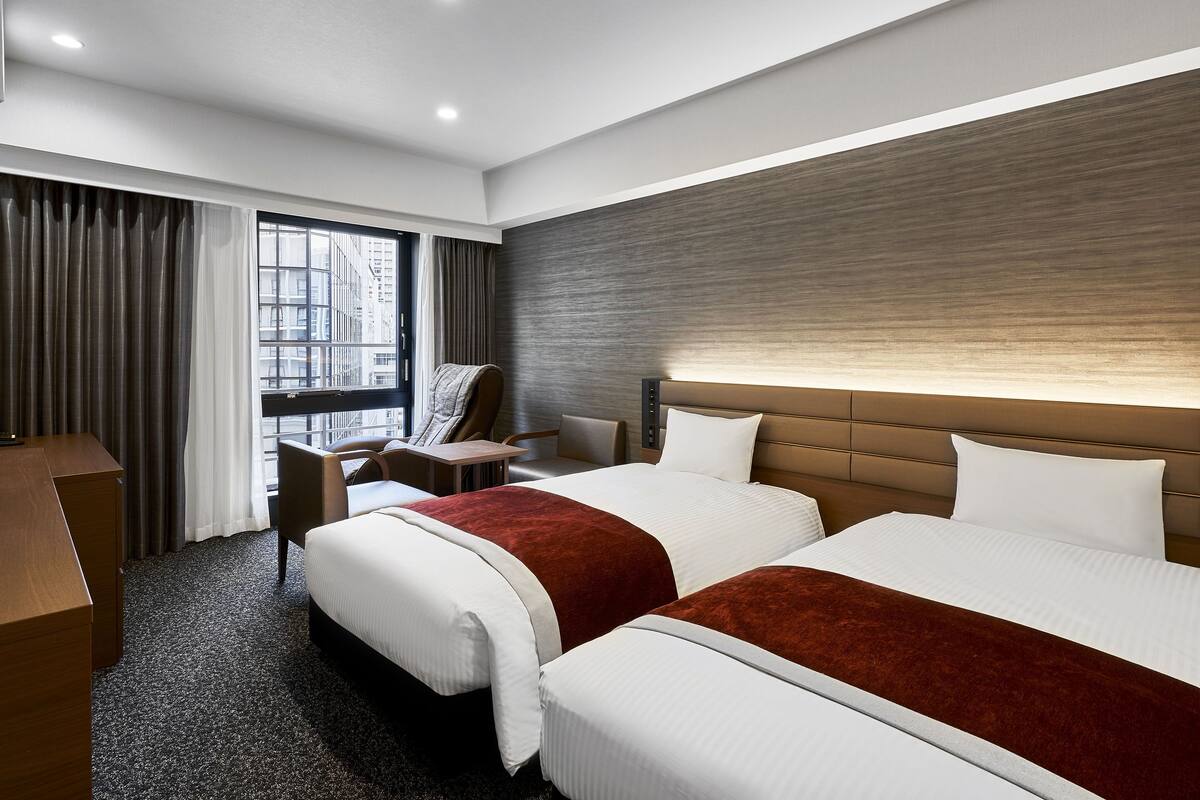 Une chambre d’hôtel avec deux lits, un bureau, une chaise et une grande fenêtre.