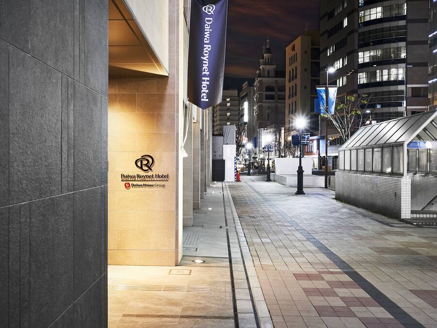 A nighttime view of a hotel entrance with a sign for Daiwa Roynet Hotel, part of the Daiwa House Group. The entrance features a tiled pathway and a modern building facade.