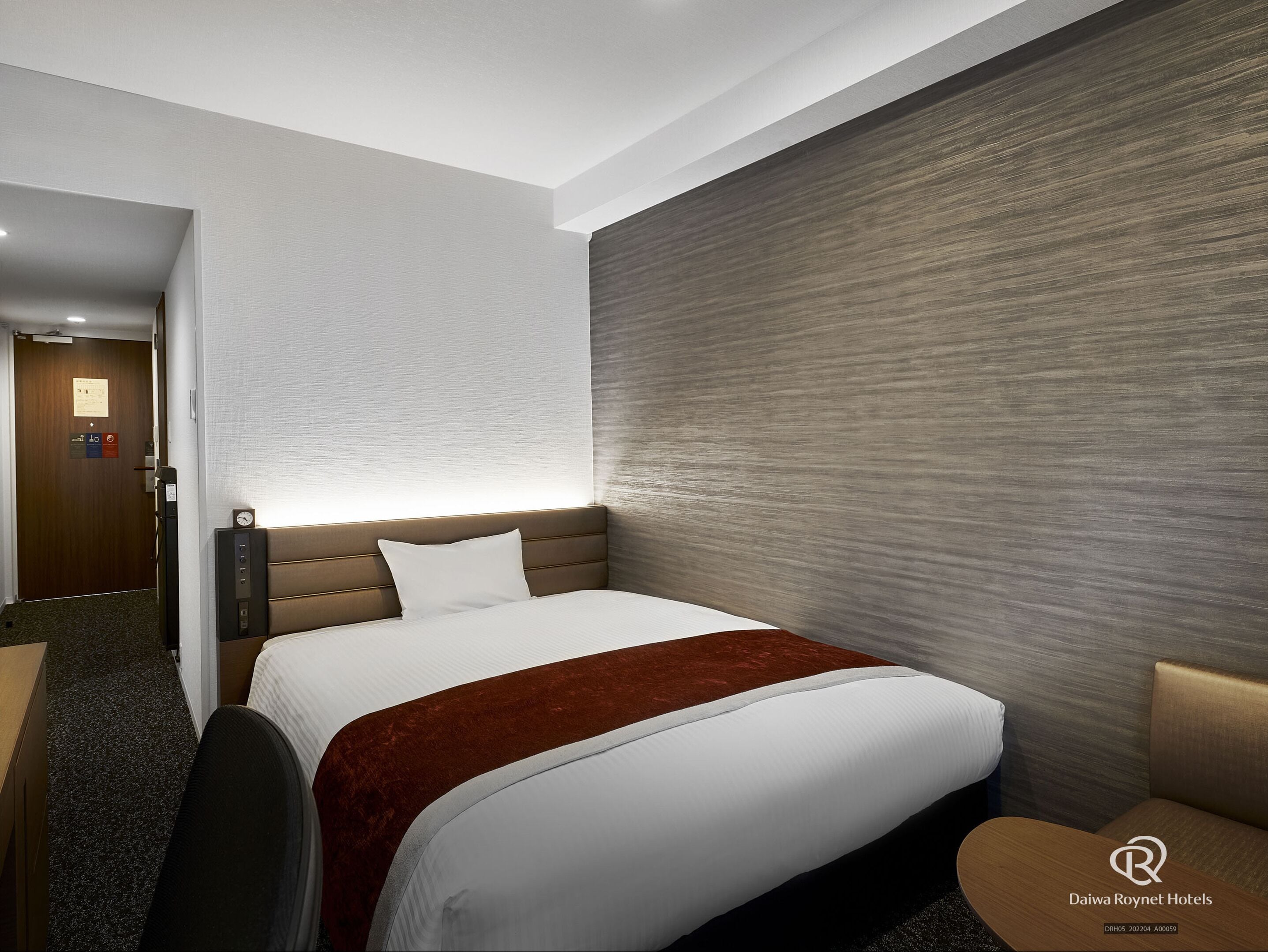 A hotel room with a bed, a desk, and a chair. There is a door with a sign above it.