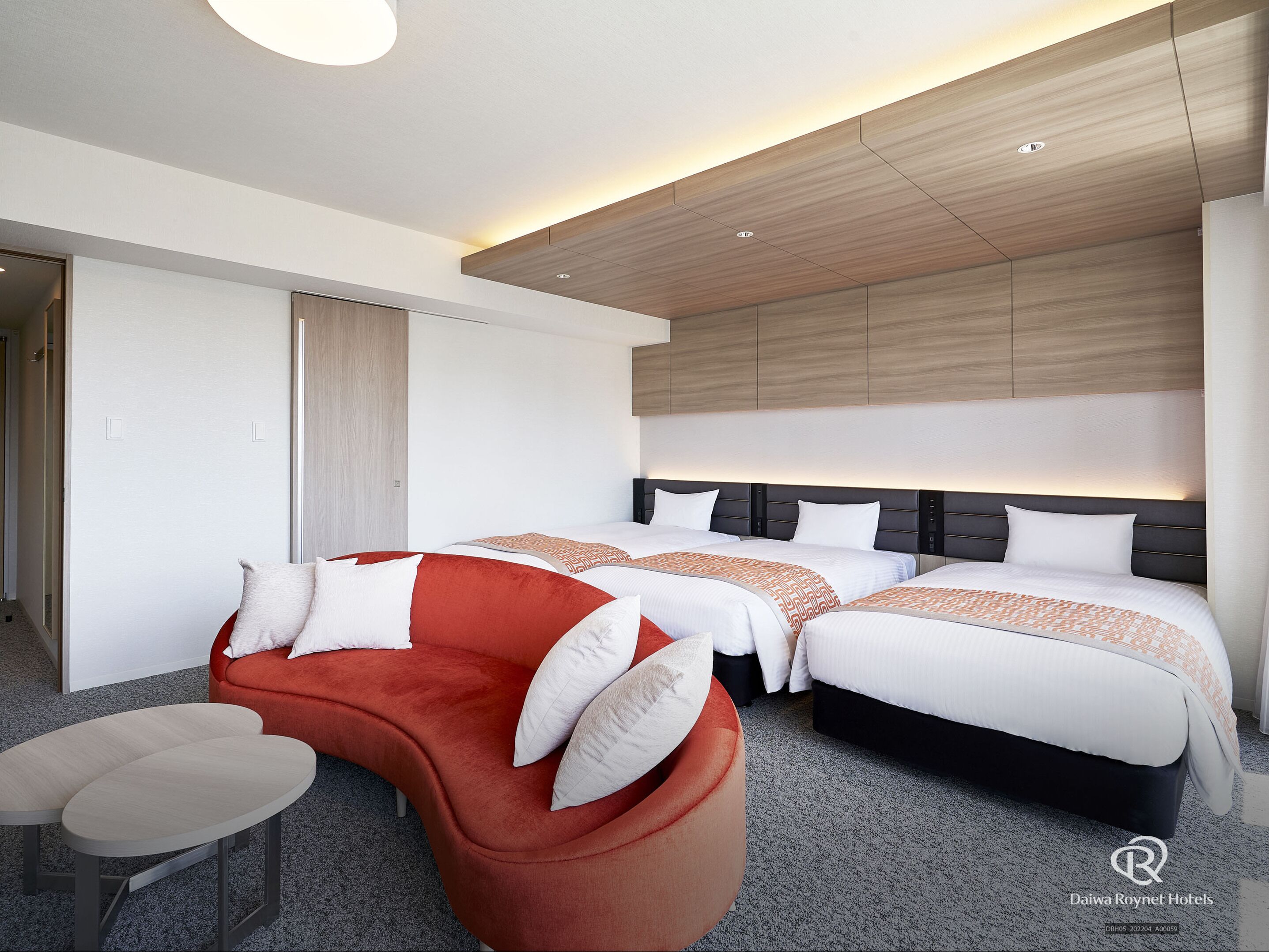 A modern hotel room with two beds, a red sofa, a small table, and a wooden ceiling.