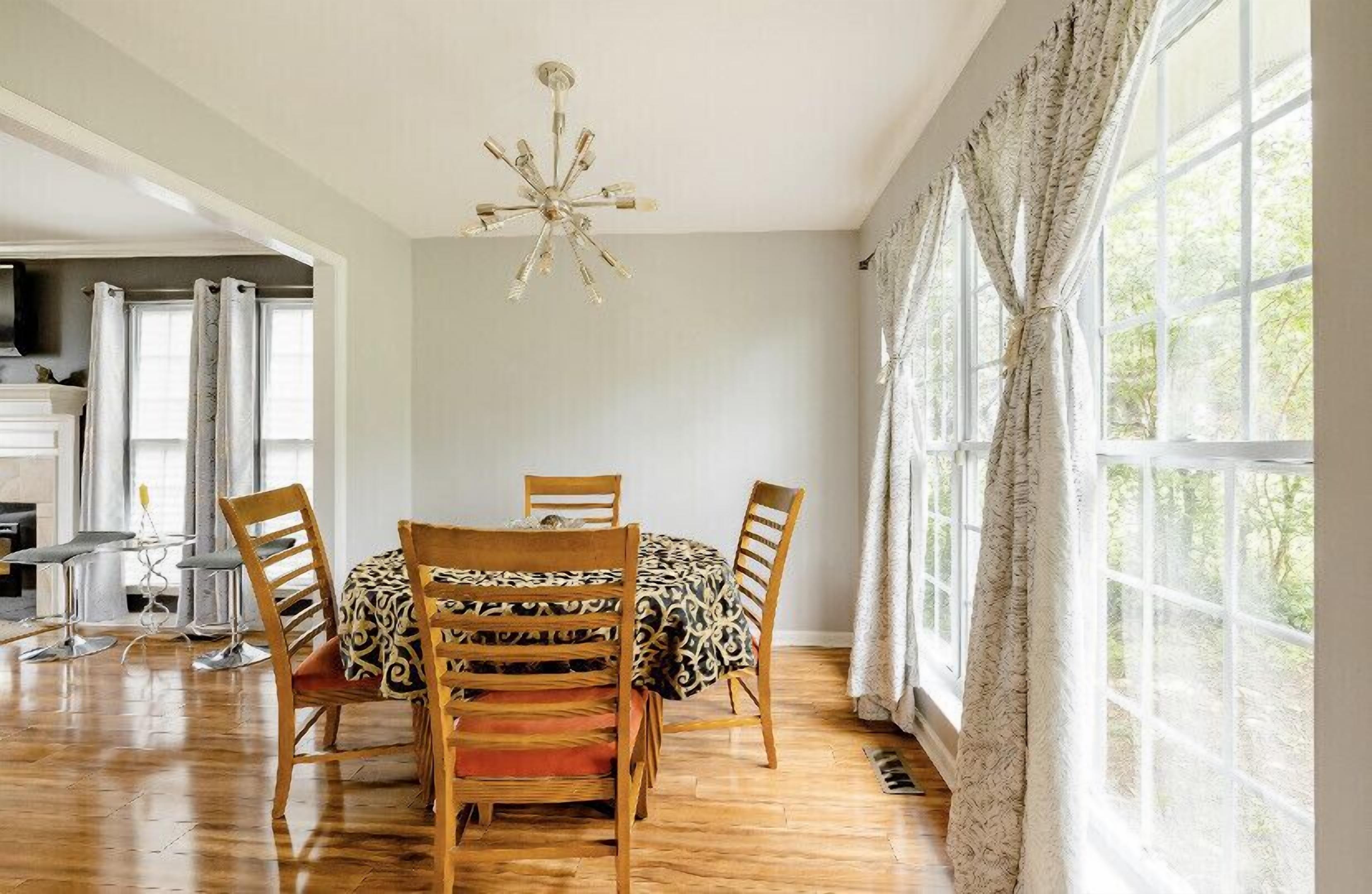 Un comedor con mesa y sillas, una araña y ventanas grandes con cortinas.