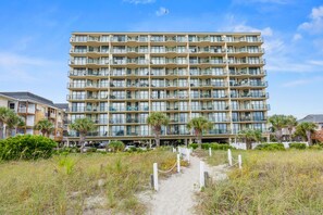 A multi-story hotel with balconies, a sandy path leading to the entrance, and palm trees.