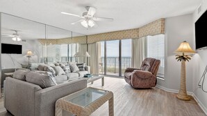 A spacious living room with a grey sofa, a brown armchair, a glass coffee table, a ceiling fan, and a large window offering a view of the beach. - Spacious family-friendly oceanfront condos w/easy beach access in North Myrtle Beach (North Myrtle Beach)