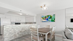 A modern kitchen with white cabinets, a tiled backsplash, and a central island. There is a dining table with chairs and a gray sofa in the living area. - Spacious family-friendly oceanfront condos w/easy beach access in North Myrtle Beach (North Myrtle Beach)