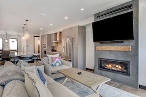 A modern living room with a fireplace, a flat-screen TV, and a dining area with a kitchen in the background. - Downtown WP River Front Villa 236  I Private Hot Tub I Views I Game Room I Discounted Attractions (Winter Park)