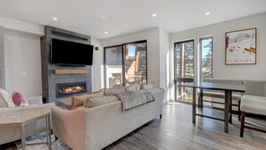 Una sala de estar moderna con chimenea, un televisor de pantalla plana, un sofá, una mesa de comedor y ventanas amplias con vista a árboles y un edificio.