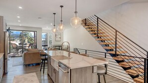 Una cocina moderna con una isla central, taburetes de bar, iluminación colgante y una escalera de madera con barandillas metálicas.