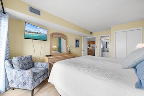 A hotel room with a bed, a chair, a dresser, a TV, a mirror, and a door leading to a kitchenette.