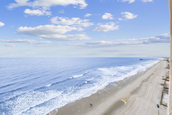 A beach with a long stretch of sand, a pier extending into the water, and a few people walking along the shore.