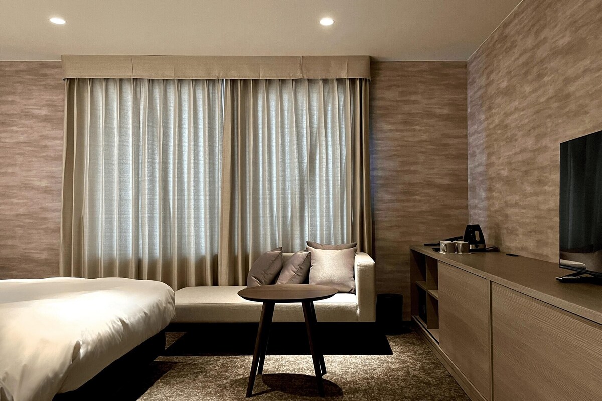 A hotel room with a bed, a sofa, a small table, a TV, and a minibar.