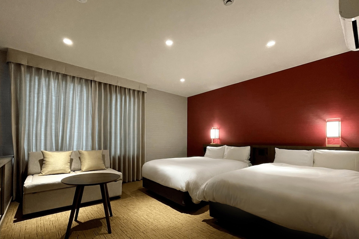 Habitación de hotel con dos camas, un sofá, una mesita y una pared decorada en rojo.