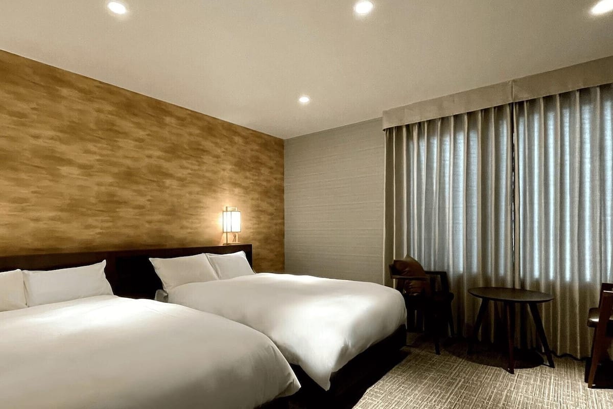 A hotel room with two beds, a small table, a chair, and a window with curtains.