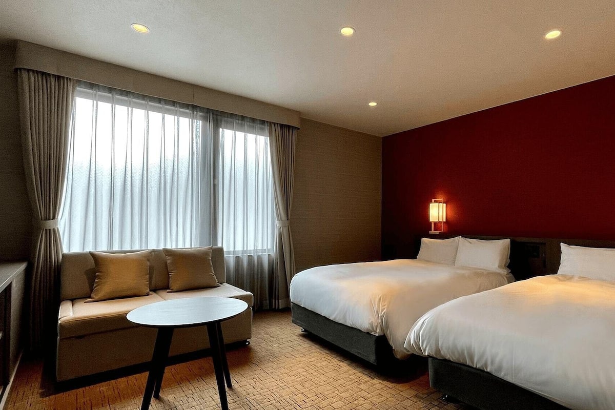 A hotel room with a sofa, two beds, a small table, and a window with curtains.