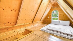 Un pequeño dormitorio de buhardilla con techo inclinado, una ventana con vistas a la vegetación y una cama con ropa blanca.