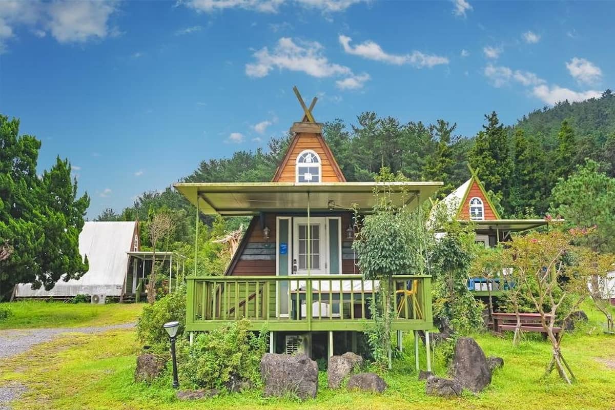Une petite cabane en bois, dotée d’un toit triangulaire et d’un porche, entourée d’une végétation luxuriante et d’arbres.