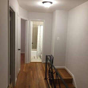 A narrow hallway with wooden flooring, white walls, and a staircase with a metal railing.