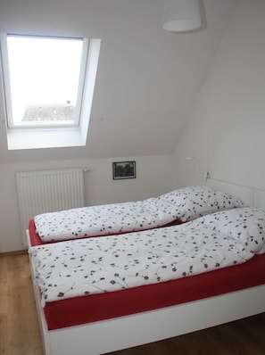 Un petit appartement d’une seule pièce, avec un plafond en pente, une fenêtre, un radiateur et un lit recouvert d’une literie à motifs floraux.