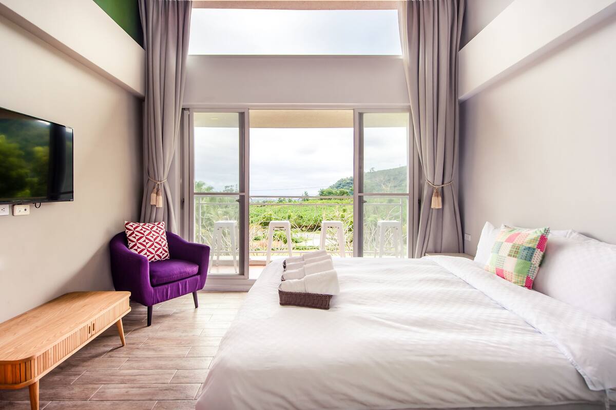 Un dormitorio con una cama grande, un sillón púrpura, un banco de madera y un televisor en la pared.