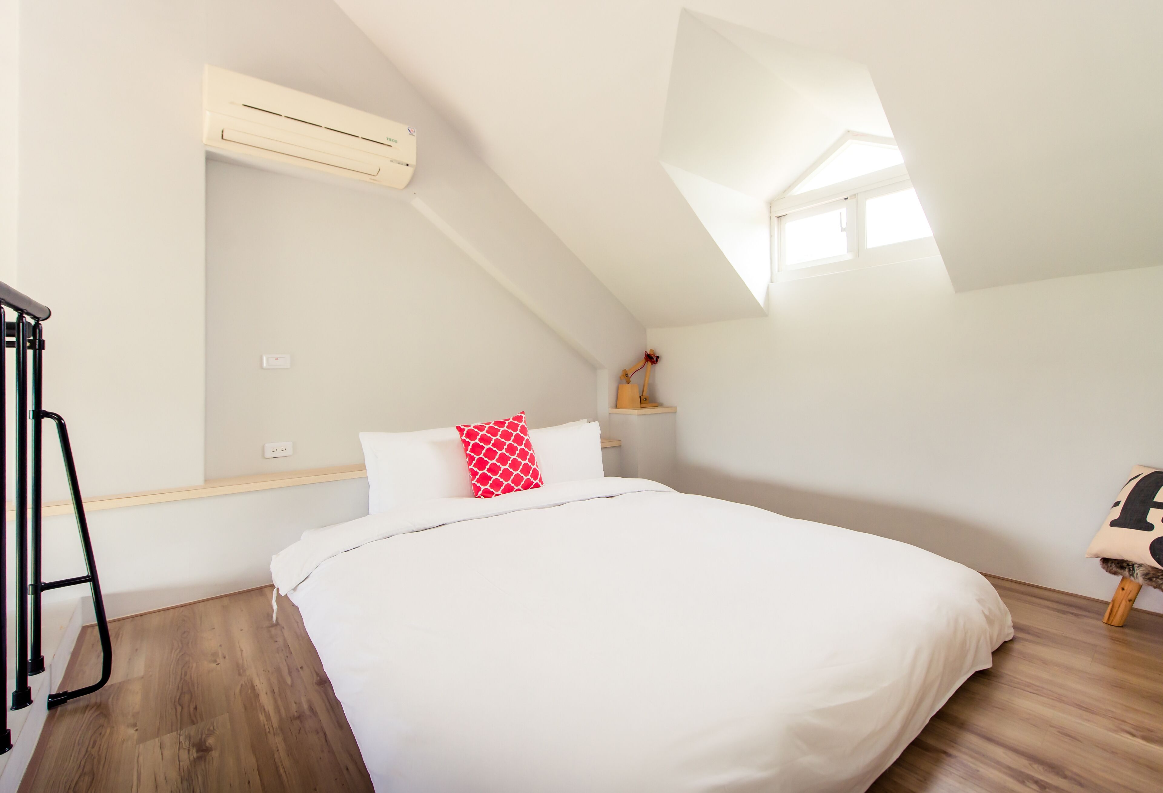 A bedroom with a bed, a red pillow, a white pillow, a wooden floor, a white wall, a white ceiling, an air conditioning unit, a window, and a small wooden shelf.