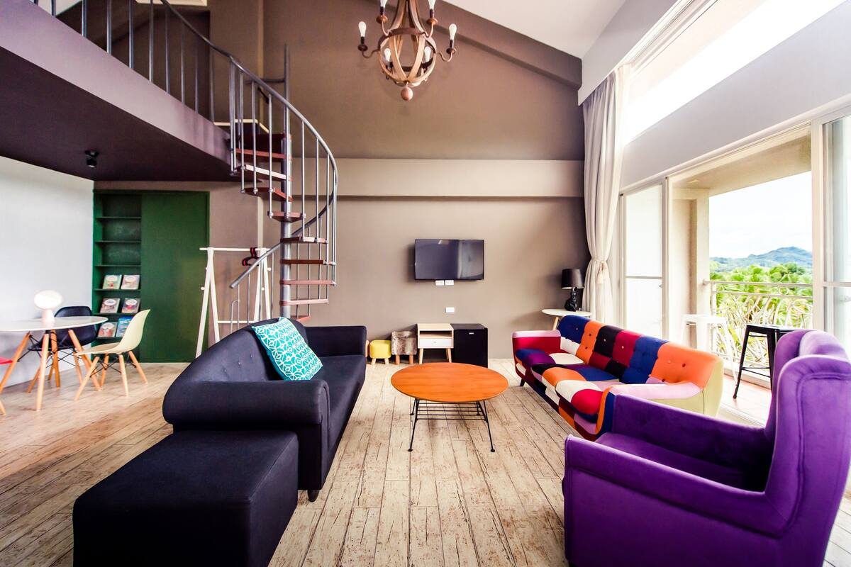Una sala moderna con un sofá negro, un sofá colorido, un sillón morado, una mesa de centro de madera y un televisor montado en la pared.