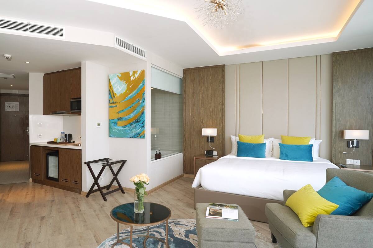 Une chambre d’hôtel moderne avec un grand lit, un canapé, une petite table ronde, un bureau et une salle de bain visible à travers une porte ouverte.