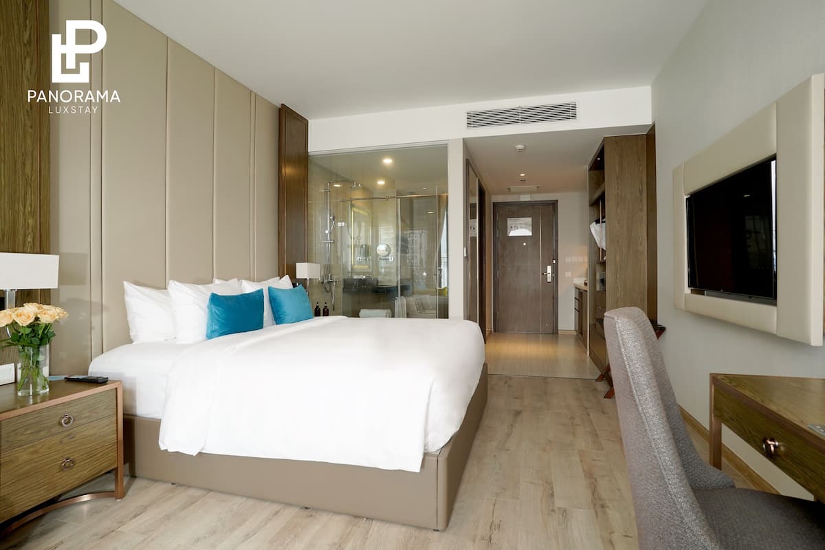 Une chambre d’hôtel moderne avec un grand lit, un téléviseur fixé au mur et une table de chevet en bois ornée d’un vase de fleurs.