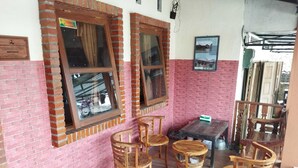 Quarto com paredes de tijolo, janelas de madeira e piso ladrilhado. Há cadeiras de madeira e uma mesa pequena.