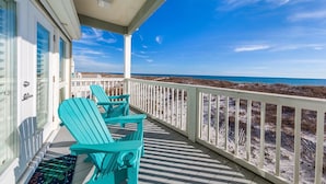 A balcony with turquoise Adirondack chairs, a white railing, and a view of the beach and ocean.