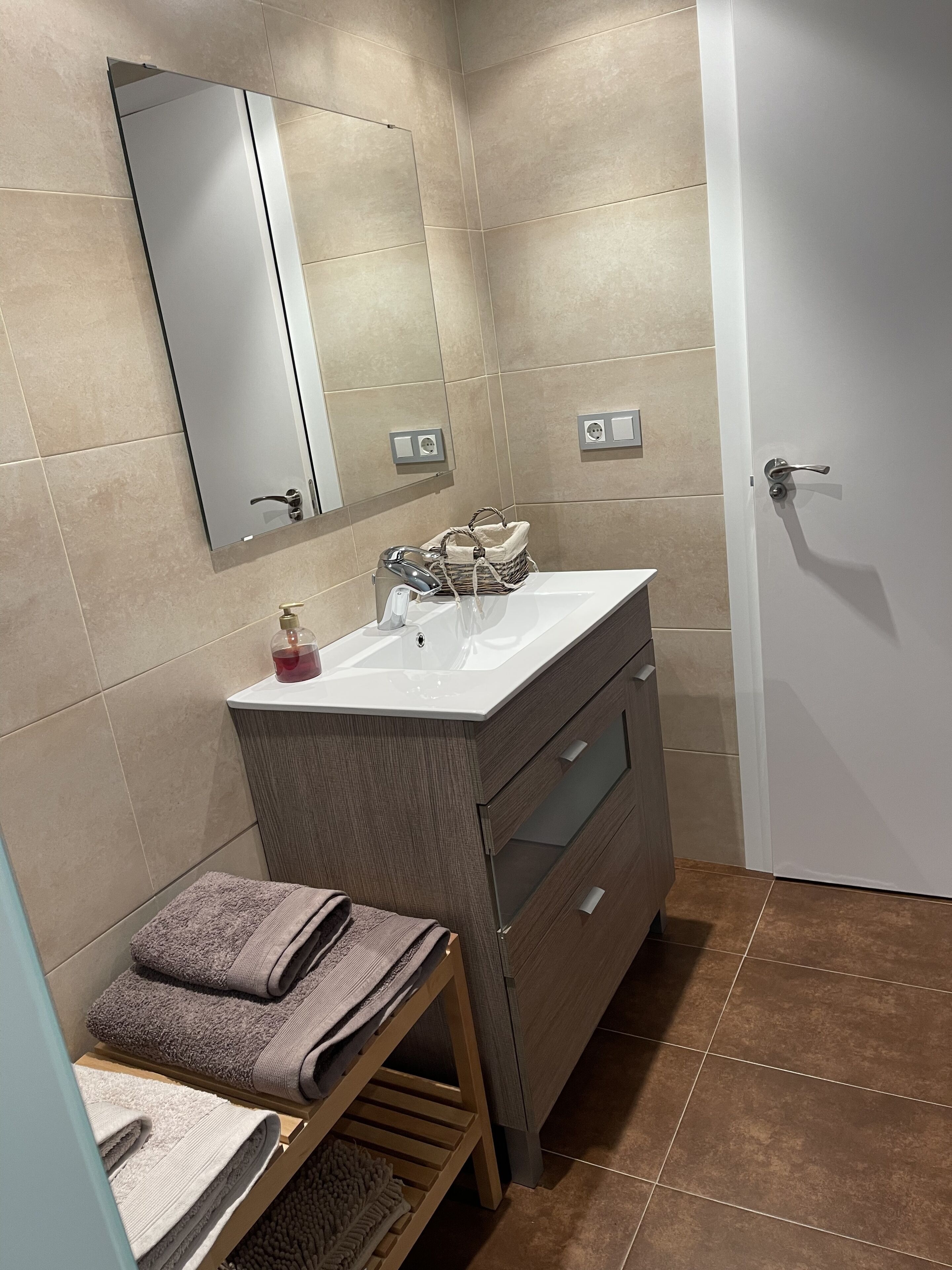A modern bathroom with a white sink, mirror, and wooden vanity. There are towels on a shelf and a door to the right.
