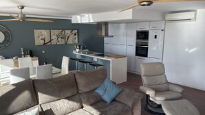 En moderne stue med sofa, lænestol og spiseområde, med et køkken i baggrunden.