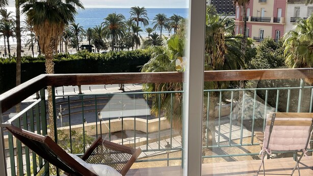 En balkon med en træstol og en loungeseng med hynder, der ud til en strand med palmer og en klar, blå himmel.