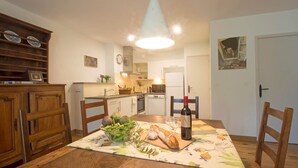 A dining area with a wooden table, chairs, a chandelier, and a kitchen in the background. - Maison des Cyprès (N°3) (Marcorignan)