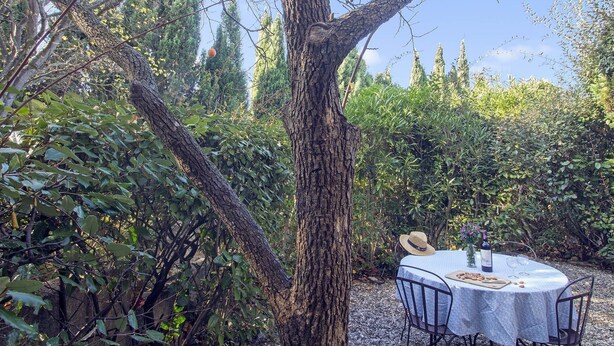 An outdoor dining area with a table set for two, surrounded by lush greenery and a large tree. - Maison des Cyprès (N°3) (Marcorignan)
